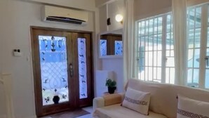 Una sala de estar con un sofá color beige, una alfombra pequeña y una planta en maceta. Hay una puerta con paneles de vidrio y un equipo de aire acondicionado arriba.