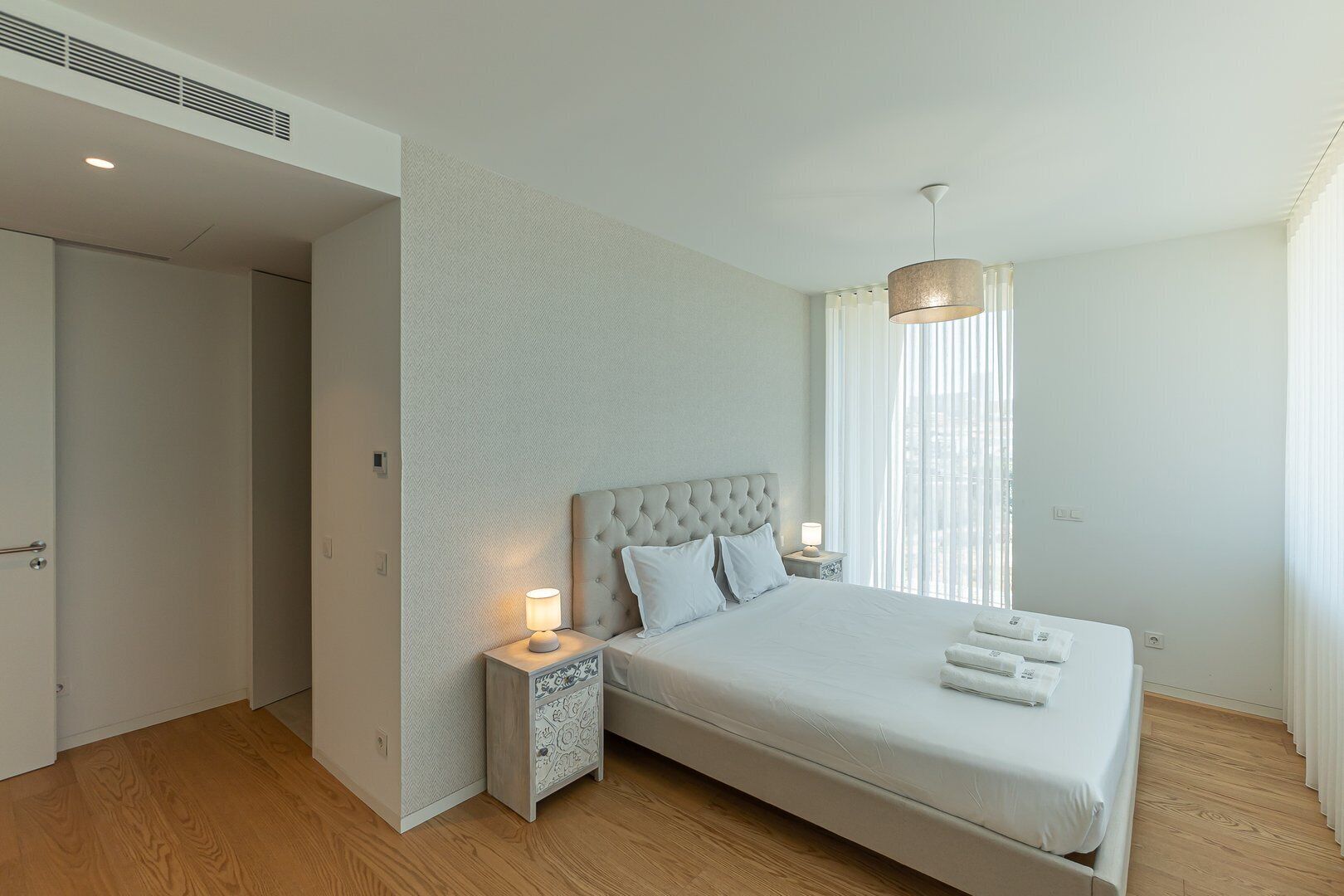 Quarto moderno com cama grande, mesa de cabeceira, abajur e vista da cidade através de cortinas translúcidas.