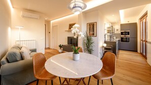 Una sala moderna con una mesa redonda de mármol, dos sillas marrones, un sofá gris, un aire acondicionado blanco, una lámpara colgante, una planta en maceta y una zona de cocina con un mueble gris y un horno.