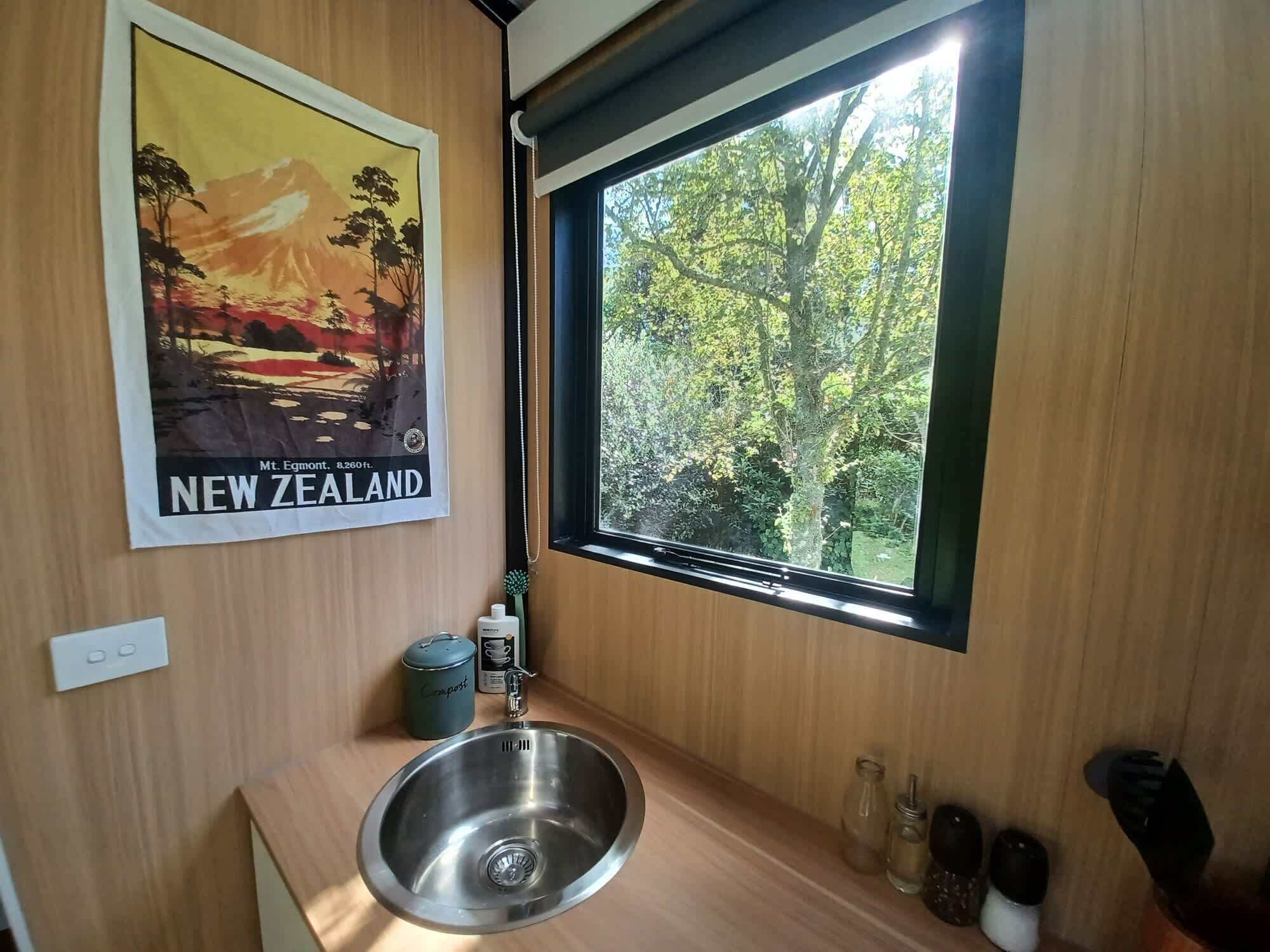 A bathroom with a stainless steel sink, a window with a view of trees, and a poster of Mt. Egmont on the wall.