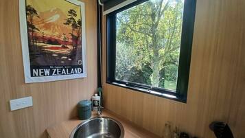 A bathroom with a stainless steel sink, a window with a view of trees, and a poster of Mt. Egmont on the wall.