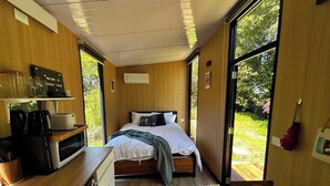 A modern bedroom with a bed, wooden walls, a small kitchenette, and large windows. - Kereru Creek by Tiny Away (Tataraimaka)