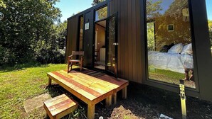 A modern, compact cabin with a wooden deck, a single chair, and a bed visible through a large glass door. - Kereru Creek by Tiny Away (Tataraimaka)