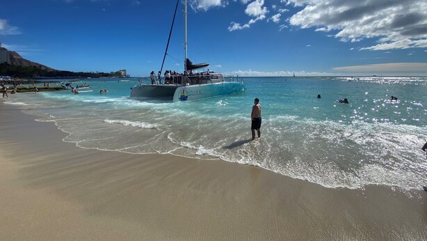 Ein Strand mit einem Segelboot namens "HAWEA" und Menschen, die schwimmen und surfen.