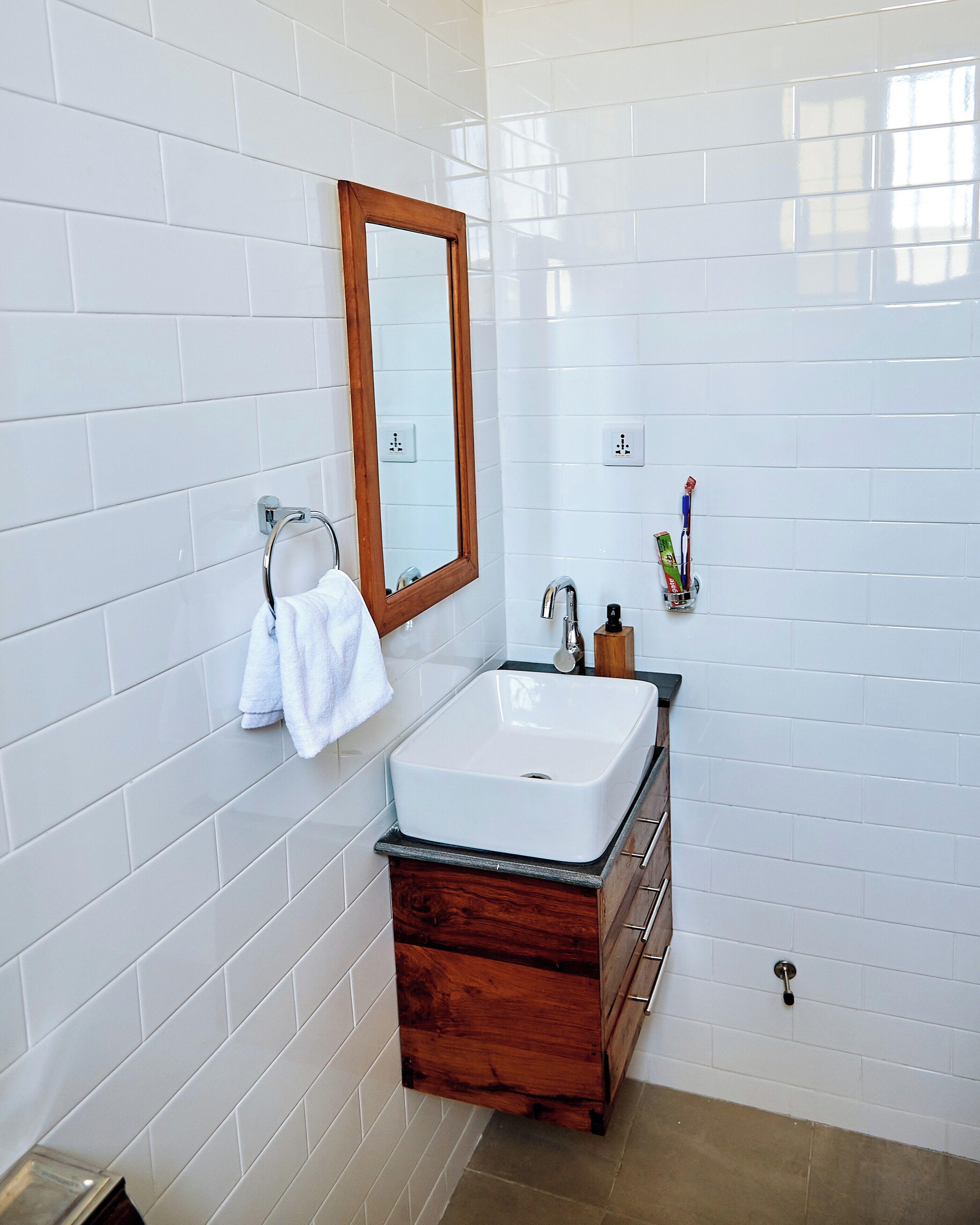 A bathroom with white tiled walls, a wooden vanity with a white sink, a mirror, and a towel rack.