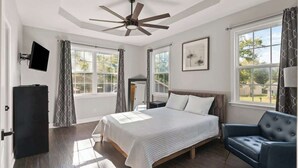 Une chambre à coucher avec un lit, un ventilateur de plafond, une télévision et une fenêtre avec des rideaux.