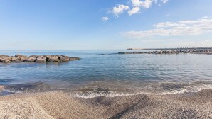 Een kiezelstrand met helder water, een rotsenrijke zeewering en een verre kustlijn met gebouwen.