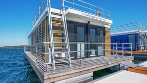 Ein schwimmendes Haus mit einer Terrasse, einer Leiter und einem Schild mit der Aufschrift 'floatinghouse.de'.