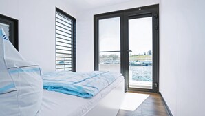 Ein Schlafzimmer mit Bett, einem Fenster mit Jalousien und einer Tür, die auf einen Balkon mit Blick auf ein Gewässer und Gebäude führt.