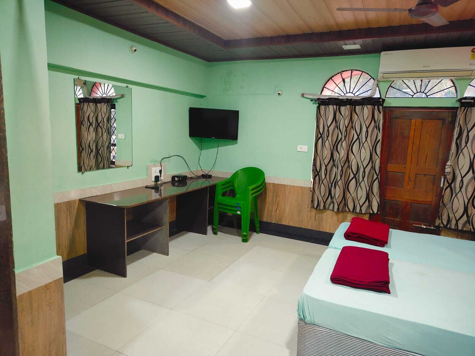 Quarto com cama, escrivaninha, cadeira, televisão e janela com cortinas.