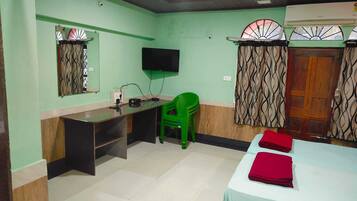 Quarto com cama, escrivaninha, cadeira, televisão e janela com cortinas.