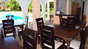A dining area with a pool view, wooden tables and chairs, and a view of the pool and greenery.