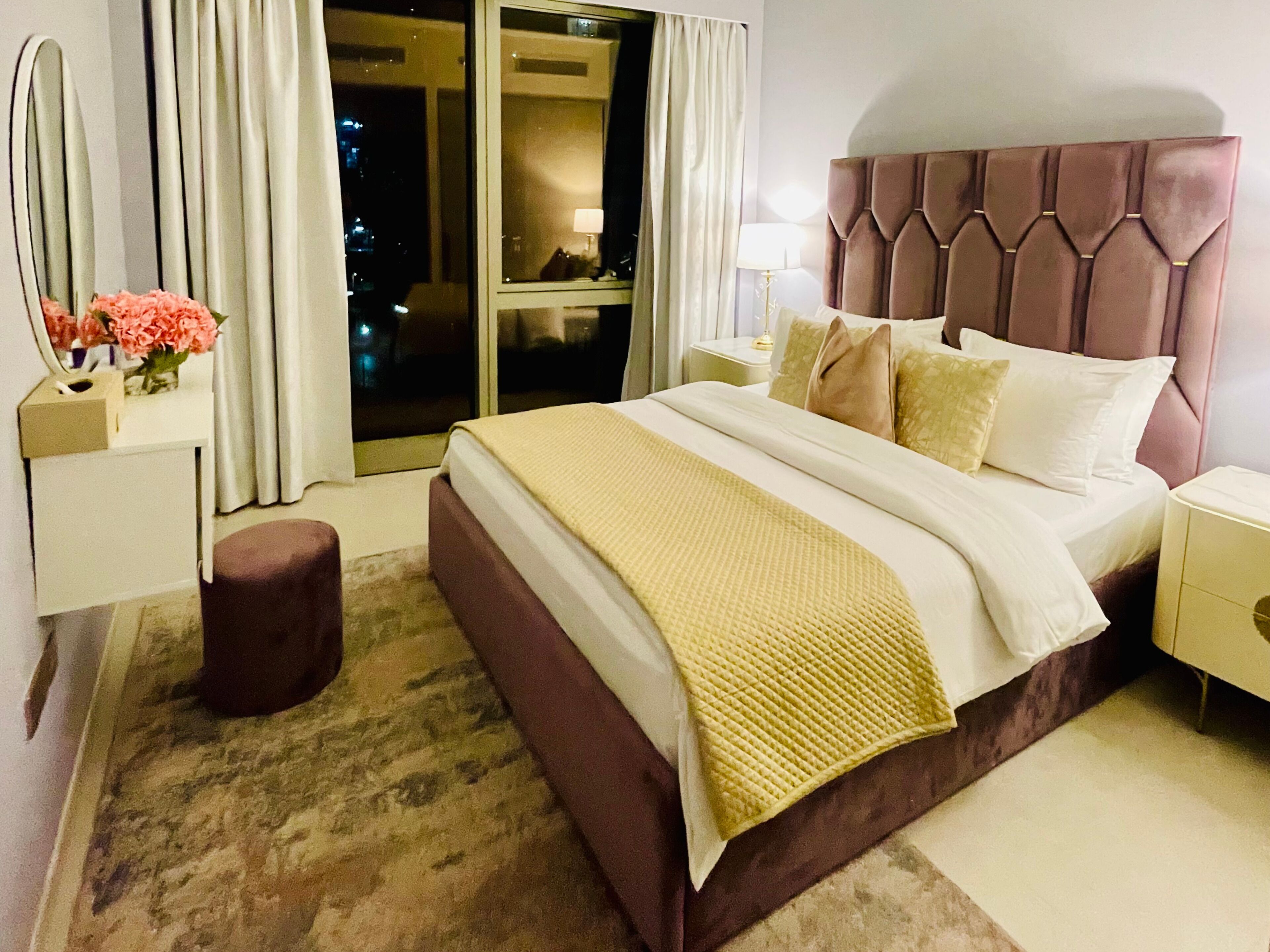 A hotel room with a large bed, a nightstand, a lamp, a mirror, and a window with curtains.