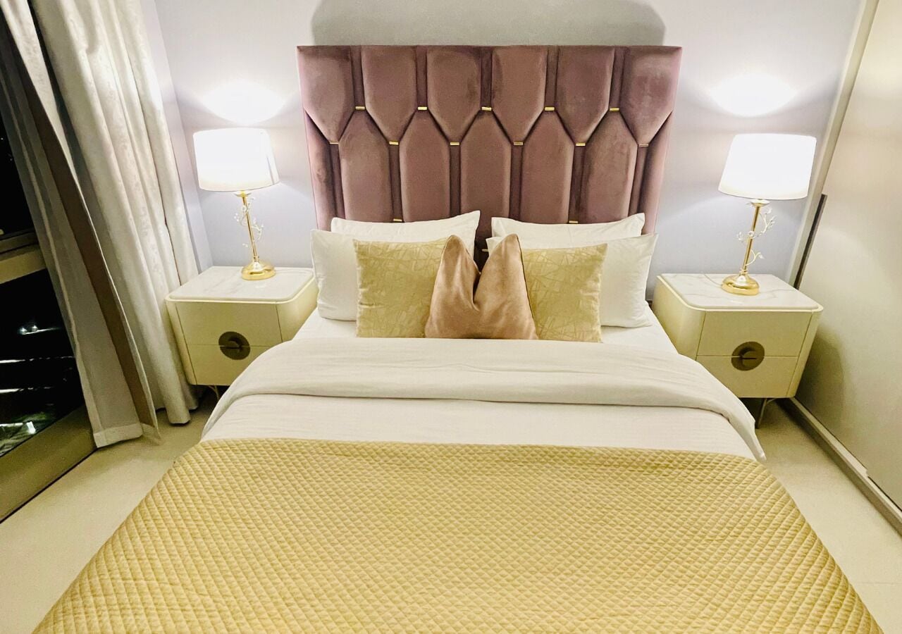 A hotel room with a tufted headboard, two bedside tables with lamps, a bed with a quilted bedspread, and a window with curtains.