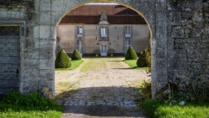 Un portail en pierre en forme d’arc donne accès à un bâtiment à la façade symétrique, doté d’une entrée centrale et d’une cour pavée bordée de haies taillées.