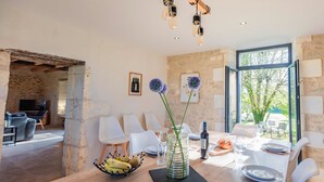 Un coin repas avec une table en bois, des chaises blanches, un vase rempli de fleurs et un saladier de fruits.