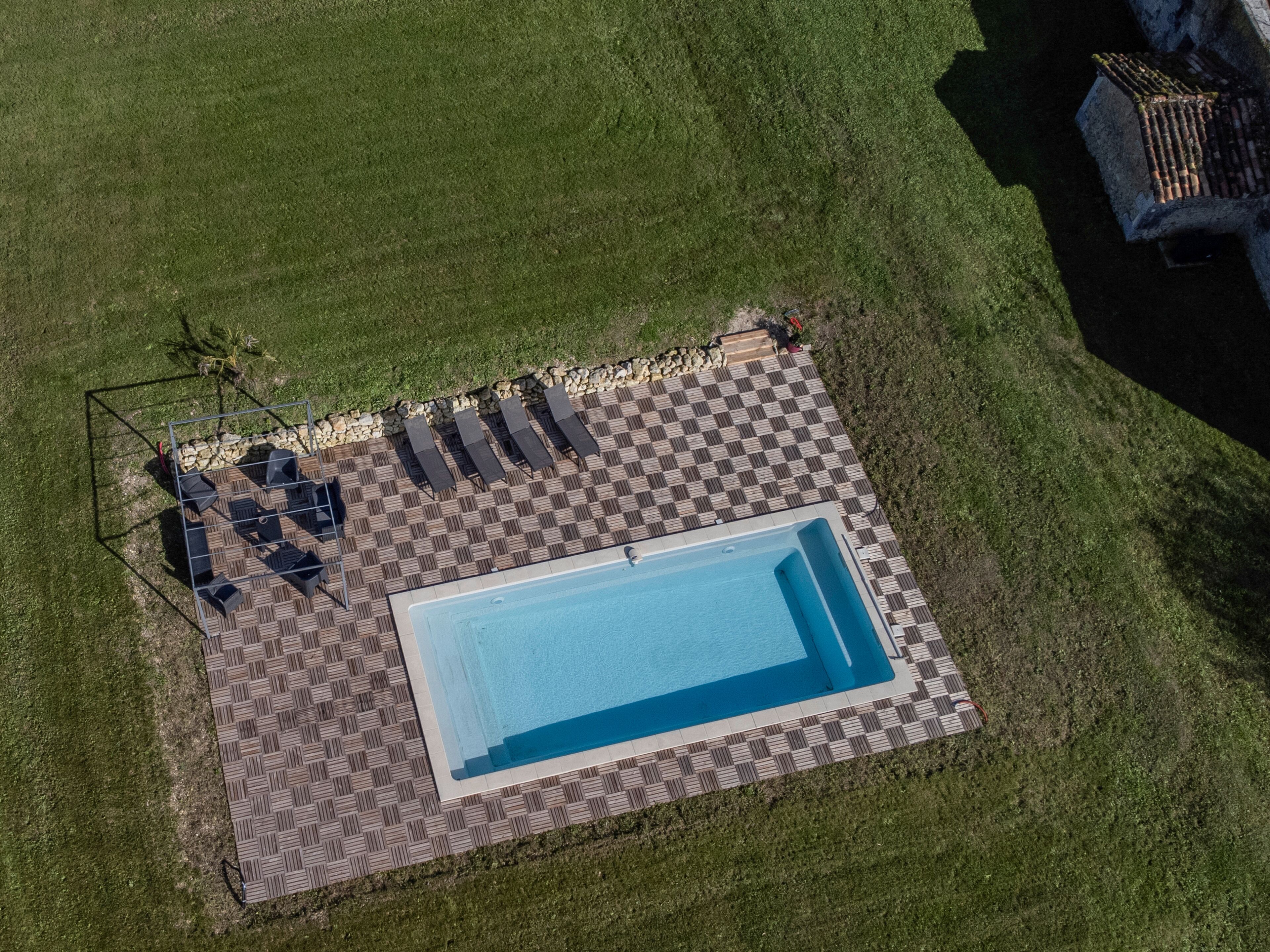 An aerial view of a rectangular swimming pool with a checkered deck, surrounded by a grassy area and a stone wall.