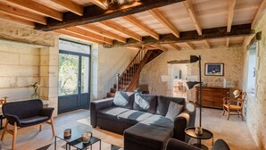 Un intérieur rustique avec des poutres apparentes, un mur en pierre, un escalier, un canapé, une table basse et un coin repas.
