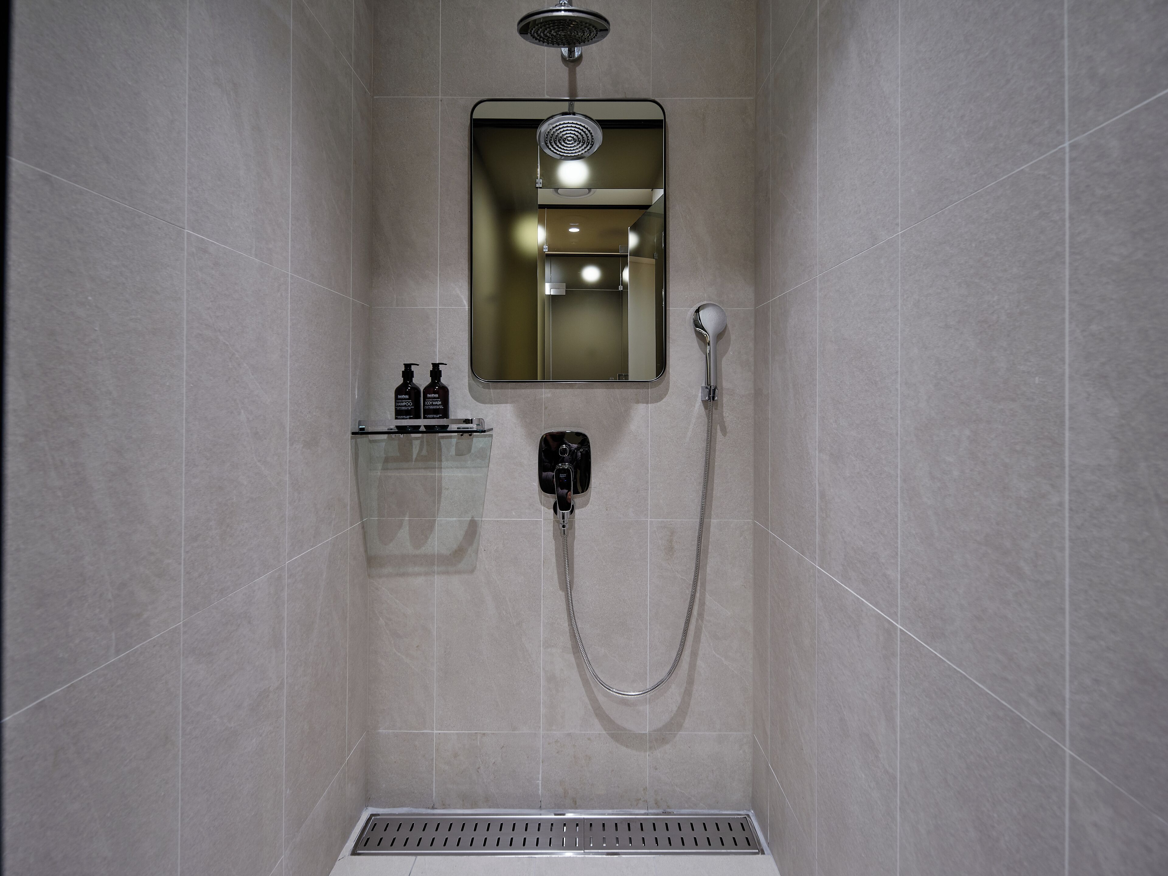 Une douche équipée d’un pommeau de douche à main, d’un miroir fixé au mur et d’une étagère pour les articles de toilette.