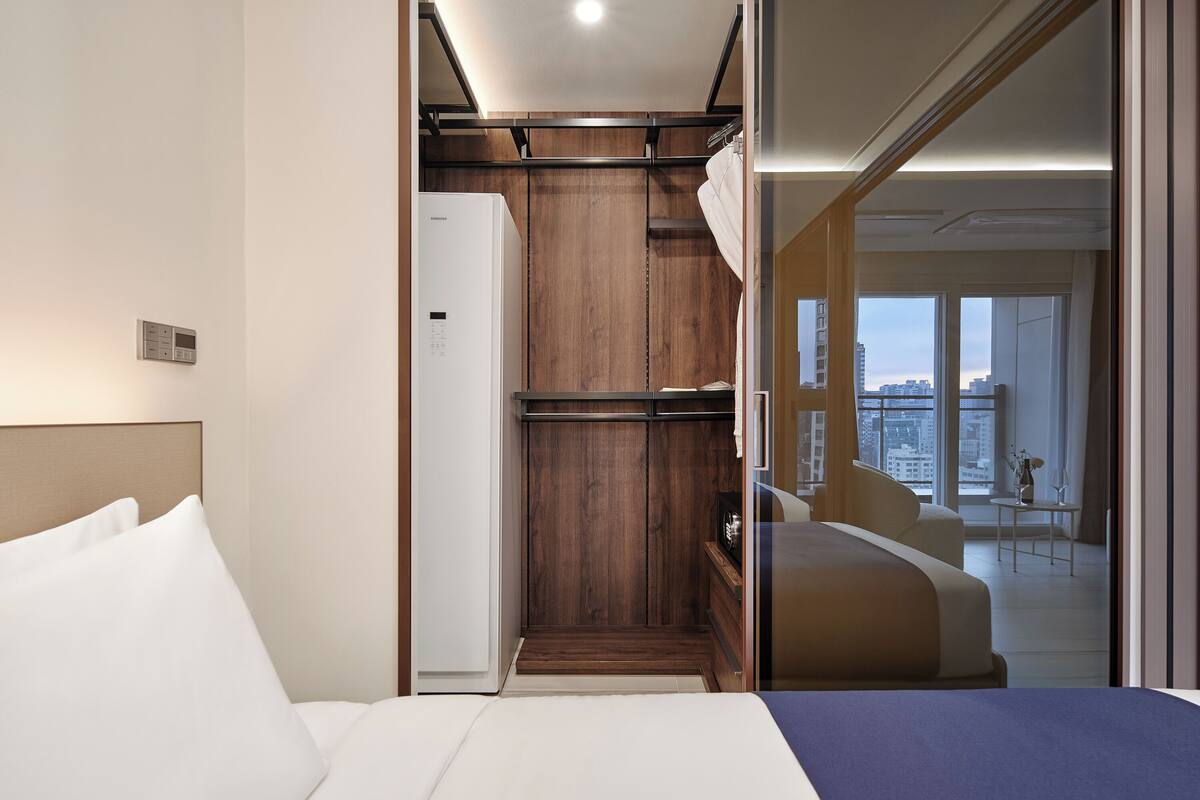 A modern hotel room with a bed, a wardrobe, and a view of the city through large windows.
