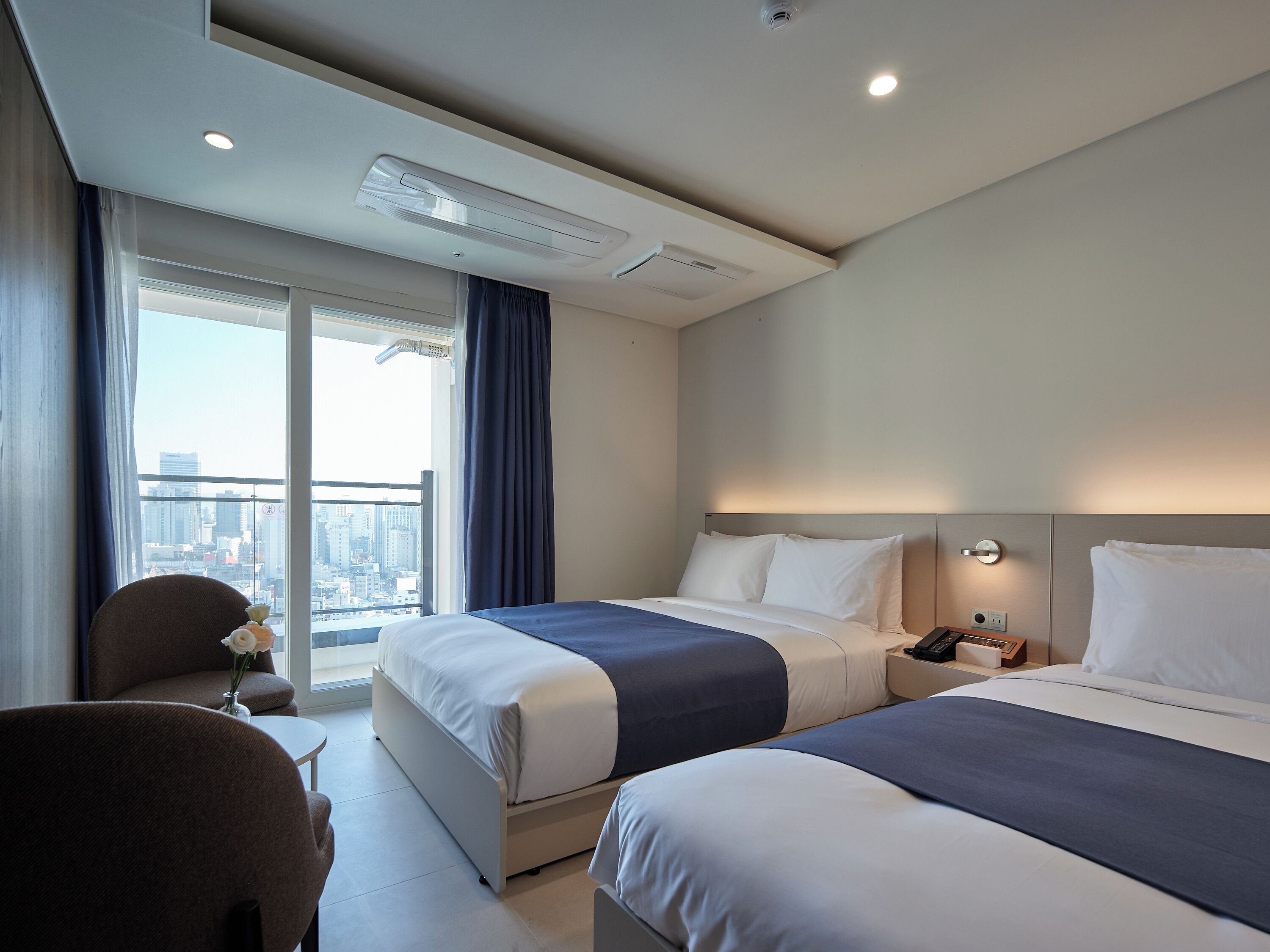 A hotel room with two beds, a desk, and a chair. There is a window with a view of the city.