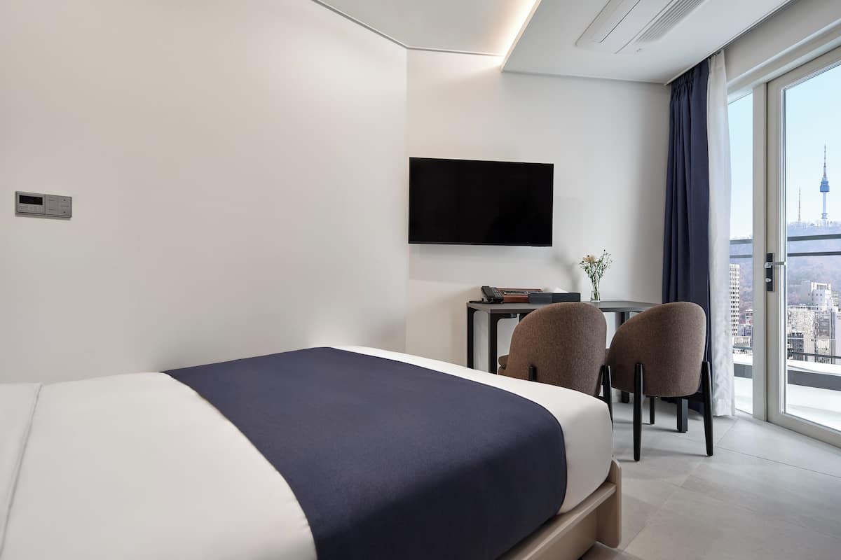 A modern hotel room with a bed, a desk, two chairs, a flat-screen TV, and a city view.