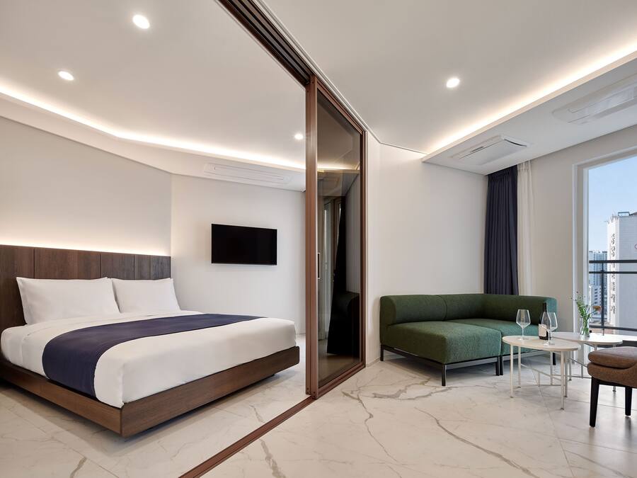 A modern hotel room with a bed, a green sofa, a small table with glasses, and a TV on the wall.