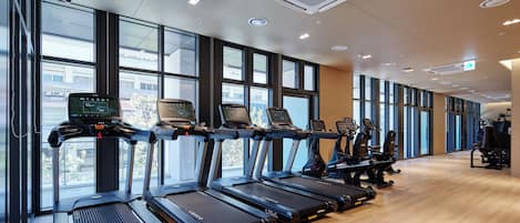 A modern gym with treadmills aligned against large windows, offering views of the city.