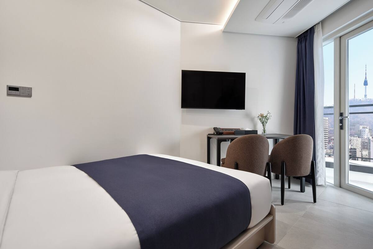 A modern hotel room with a bed, a desk, two chairs, a flat-screen TV, and a city view.
