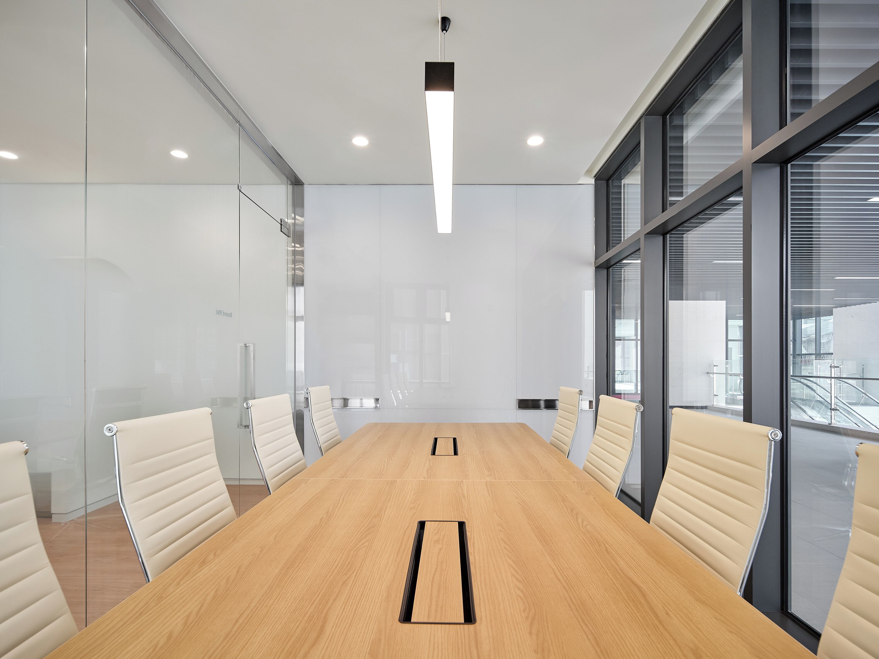 A conference room with a long wooden table, white leather chairs, glass walls, and large windows.