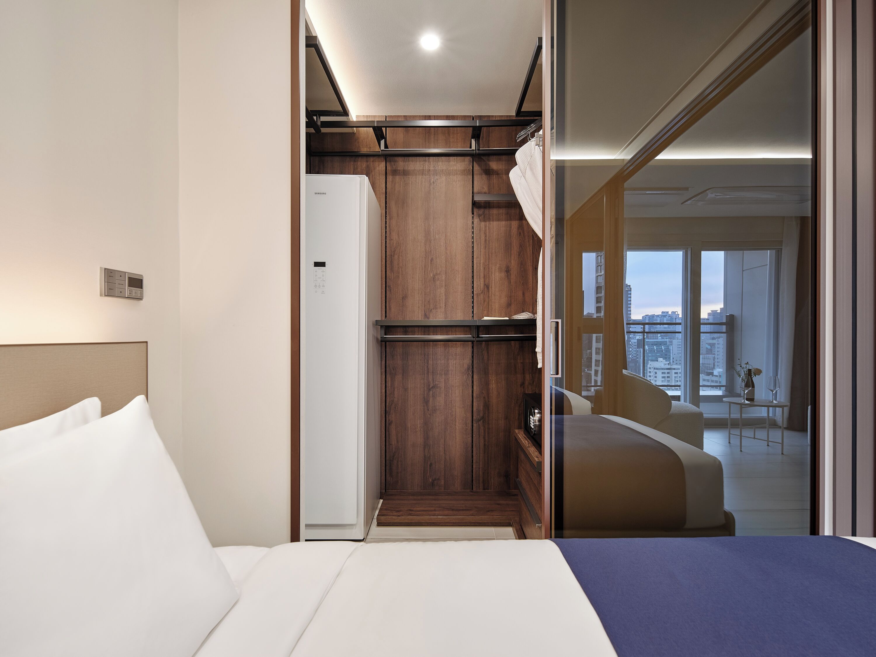 A modern hotel room with a bed, a wardrobe, and a view of the city through large windows.