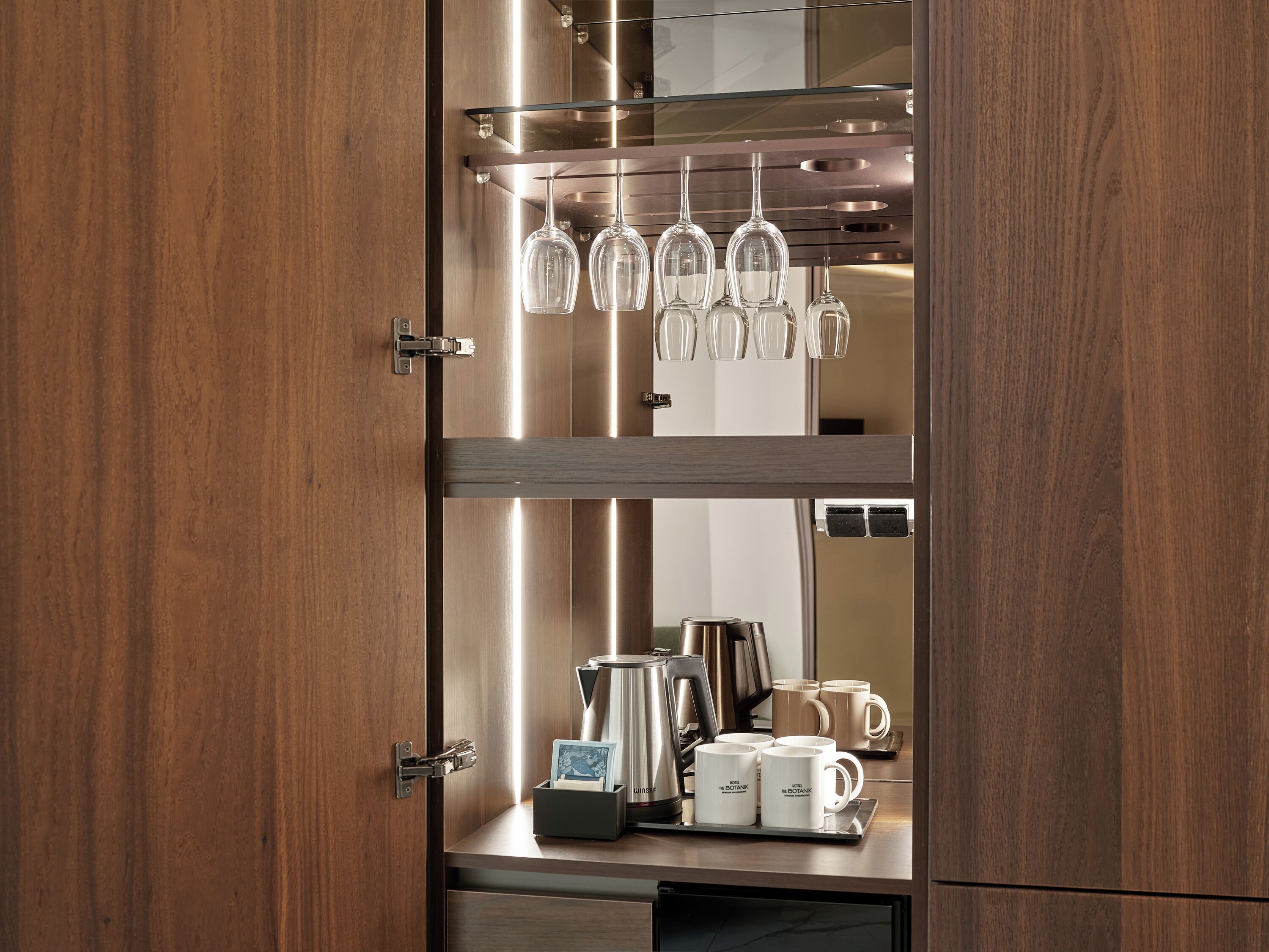 A wooden cabinet with a glass shelf holding wine glasses and a coffee maker on the lower shelf.