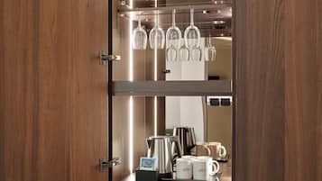A wooden cabinet with a glass shelf holding wine glasses and a coffee maker on the lower shelf.
