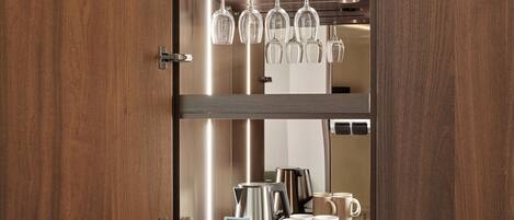 A wooden cabinet with a glass shelf holding wine glasses and a coffee maker on the lower shelf.
