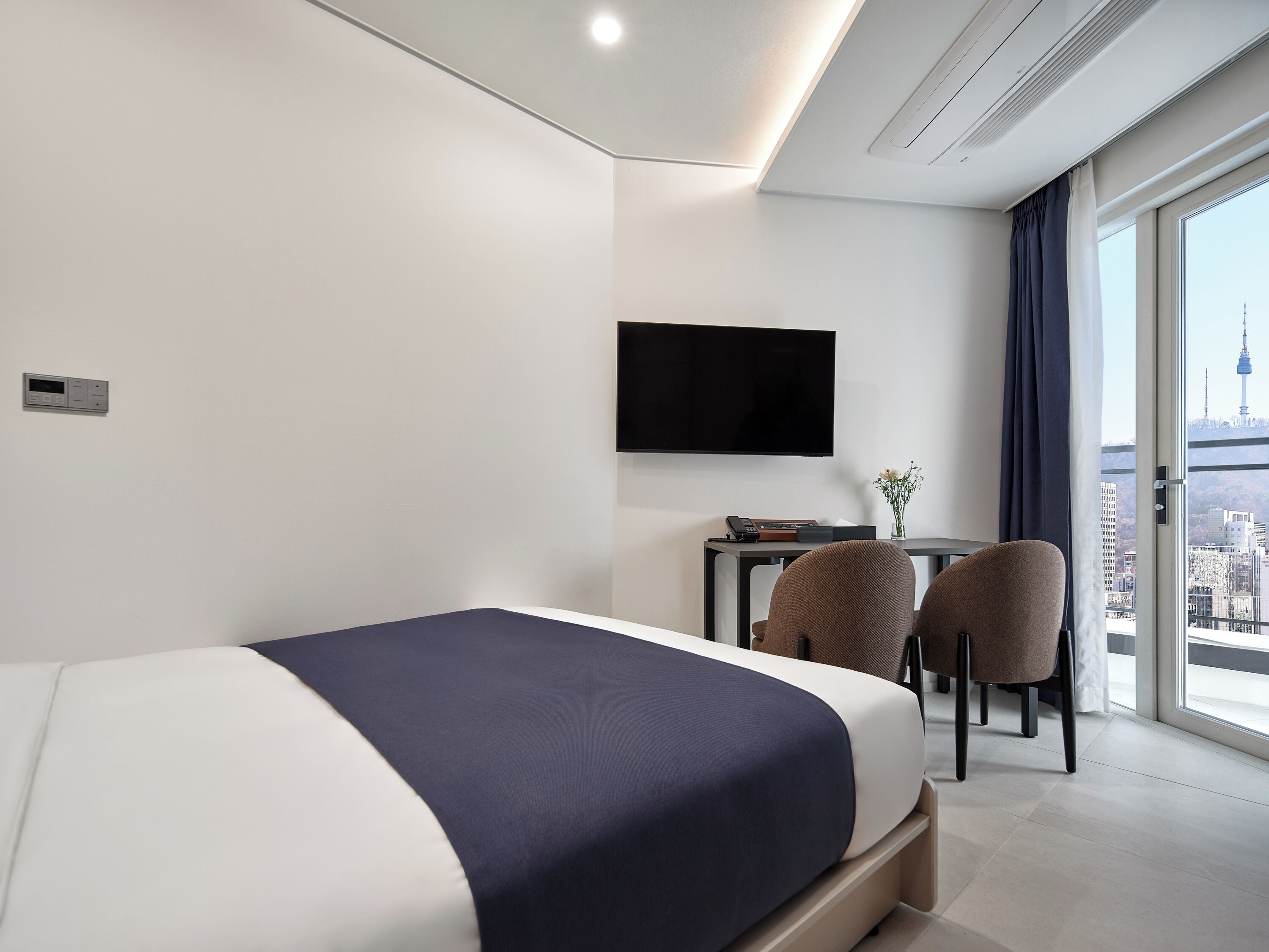 A modern hotel room with a bed, a desk, two chairs, a flat-screen TV, and a city view.