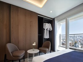 A modern hotel room with a large bed, two armchairs, a small table with a vase, and a view of the city through large windows.