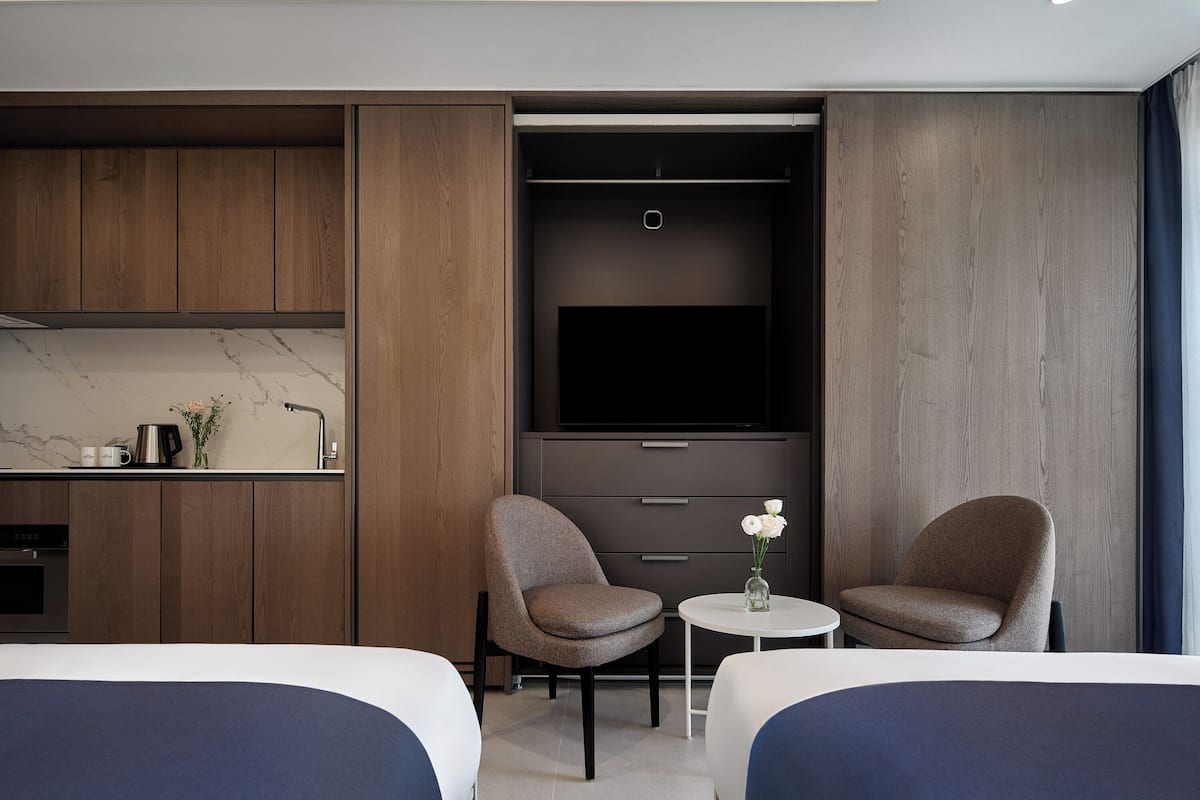 A modern hotel room with a bed, two armchairs, a small table, a TV, and a minibar.