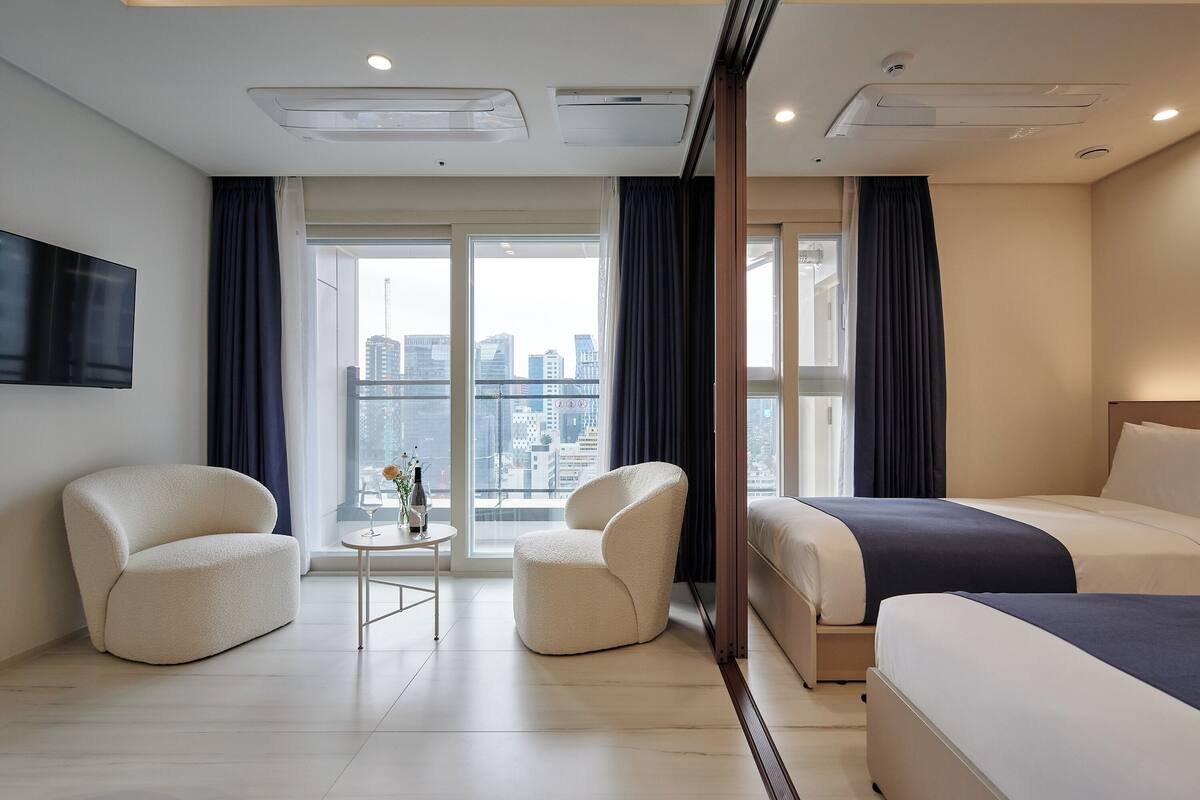A modern hotel room with a large bed, two armchairs, a small table with a vase, and a city view through large windows.