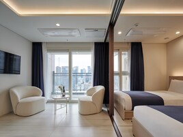 Habitación de hotel moderna con una cama grande, dos sillones, una mesita con un florero y vistas a la ciudad a través de amplias ventanas.