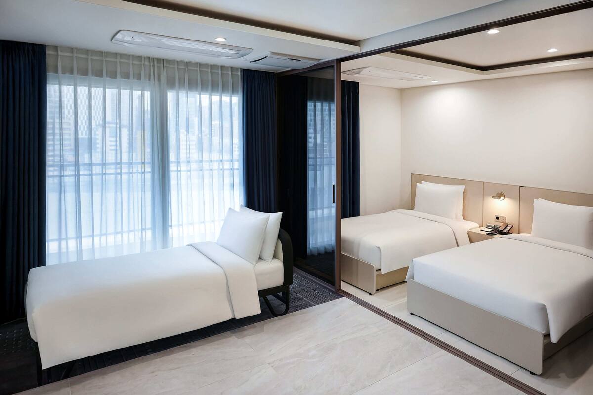 A hotel room with two beds, a large window with curtains, a bedside table, and a wall-mounted light.
