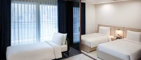A hotel room with two beds, a large window with curtains, a bedside table, and a wall-mounted light.