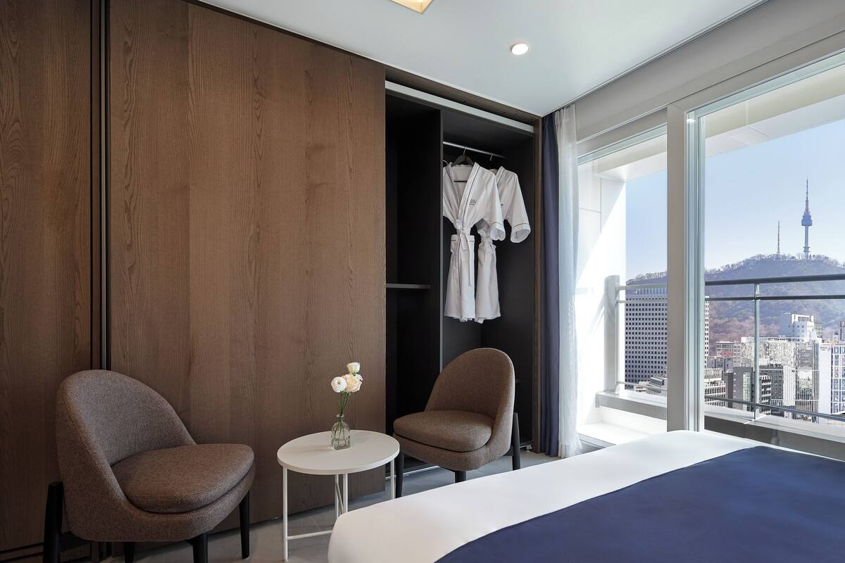 A modern hotel room with a large bed, two armchairs, a small table with a vase of flowers, and a view of the cityscape through large windows.