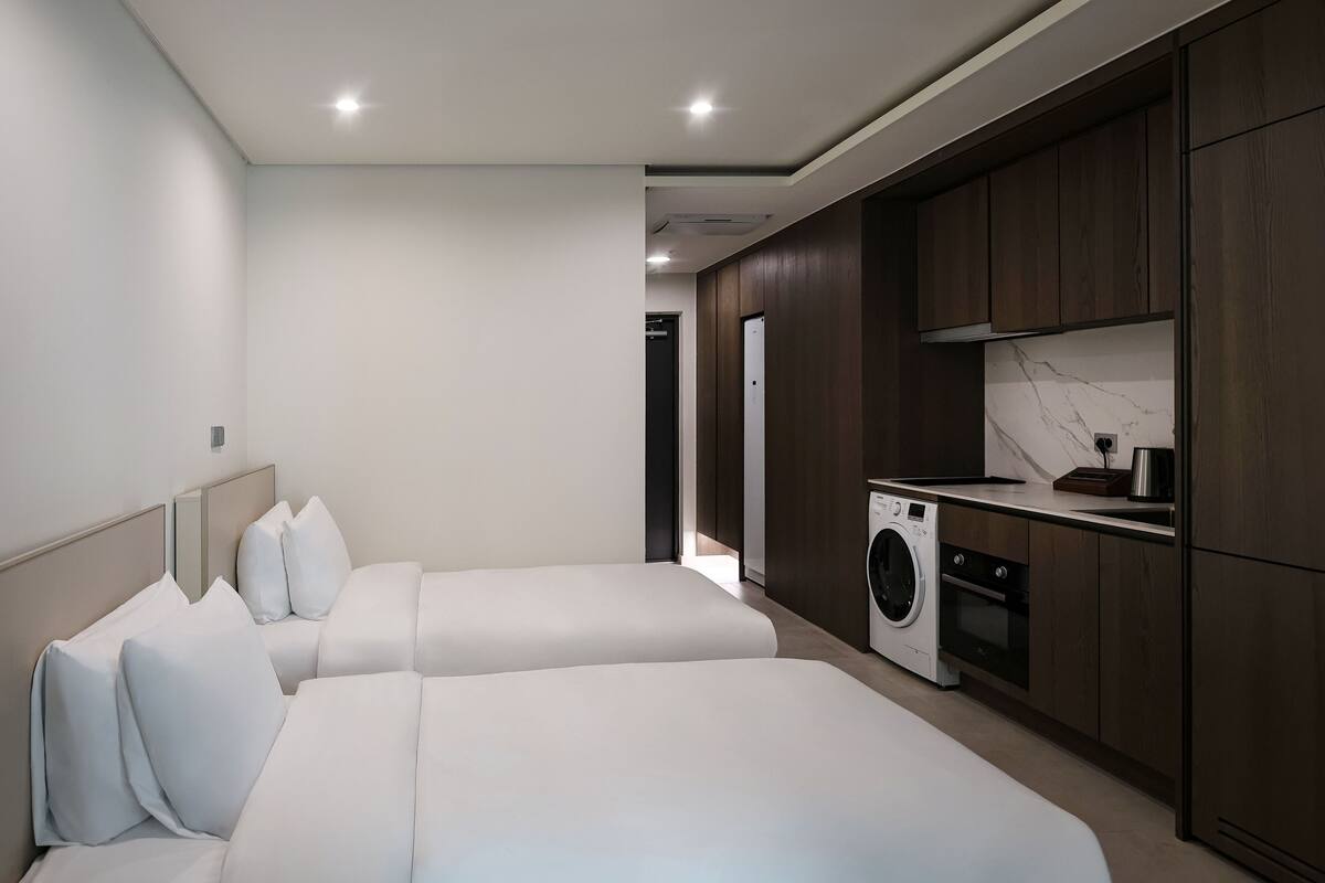 A modern hotel room with two beds, a washing machine, and a kitchenette.