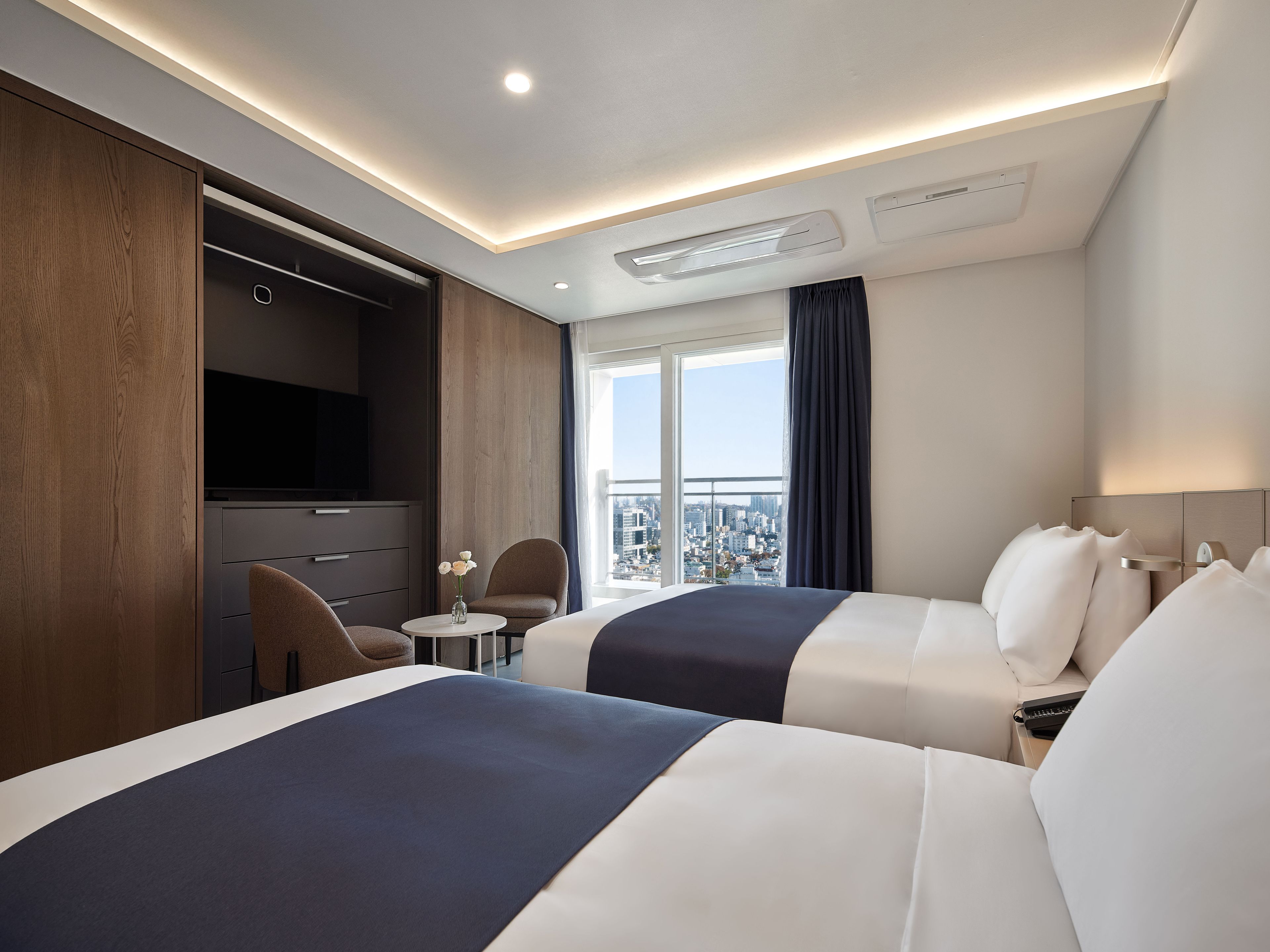 A hotel room with two beds, a TV, a small table with a vase, and a view of the city.