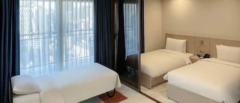 A hotel room with a single bed, a sofa bed, and a separate bed. There is a window with curtains, a bedside table with a phone, and a wall-mounted light fixture.