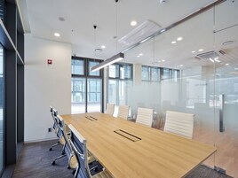 A conference room with a long wooden table, white chairs, glass partitions, and large windows.