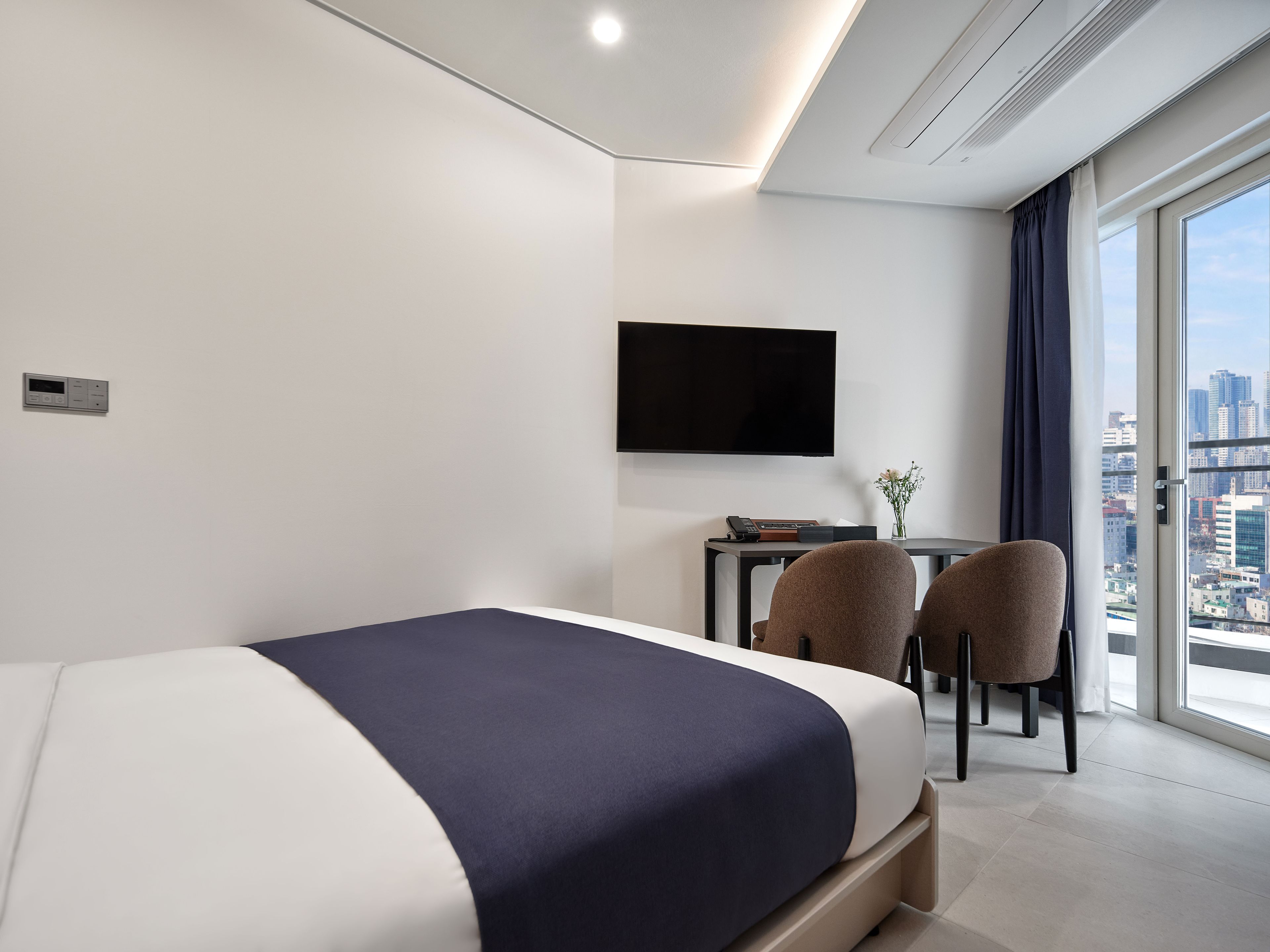 A modern hotel room with a bed, a desk, two chairs, a flat-screen TV, and a city view.