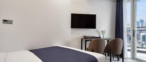 A modern hotel room with a bed, a desk, two chairs, a flat-screen TV, and a city view.