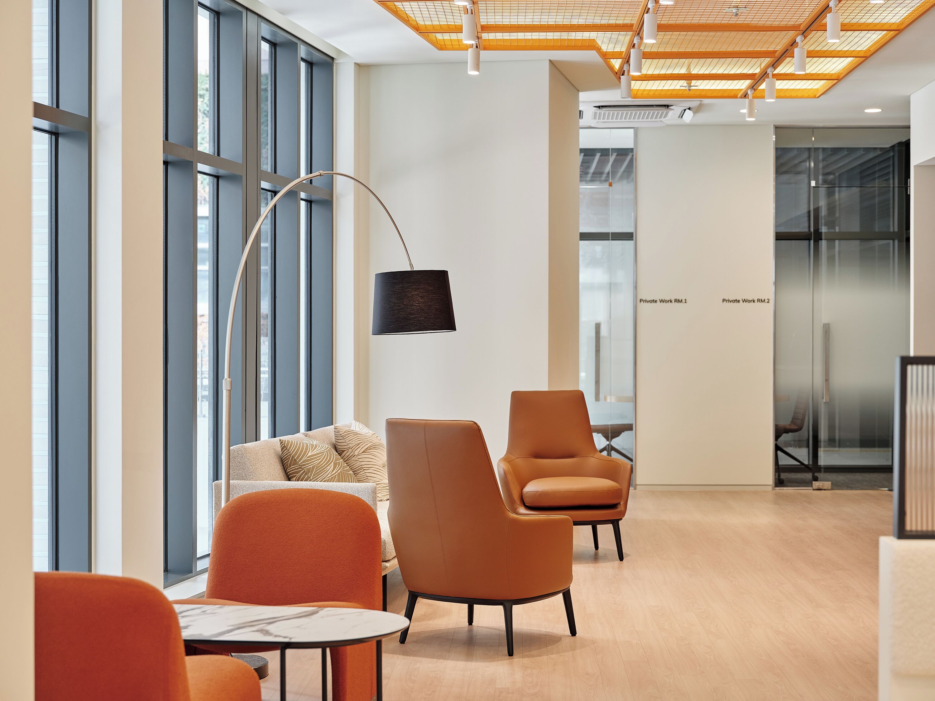 Un espacio de oficina moderno con amplios ventanales, una lámpara de techo curva y una zona de estar con sillas naranja y un sofá.