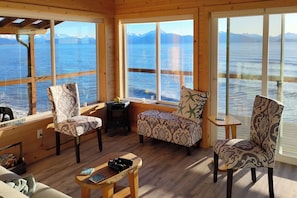 A cozy cabin with large windows overlooking a lake and mountains. There are two patterned chairs and a small table with binoculars.